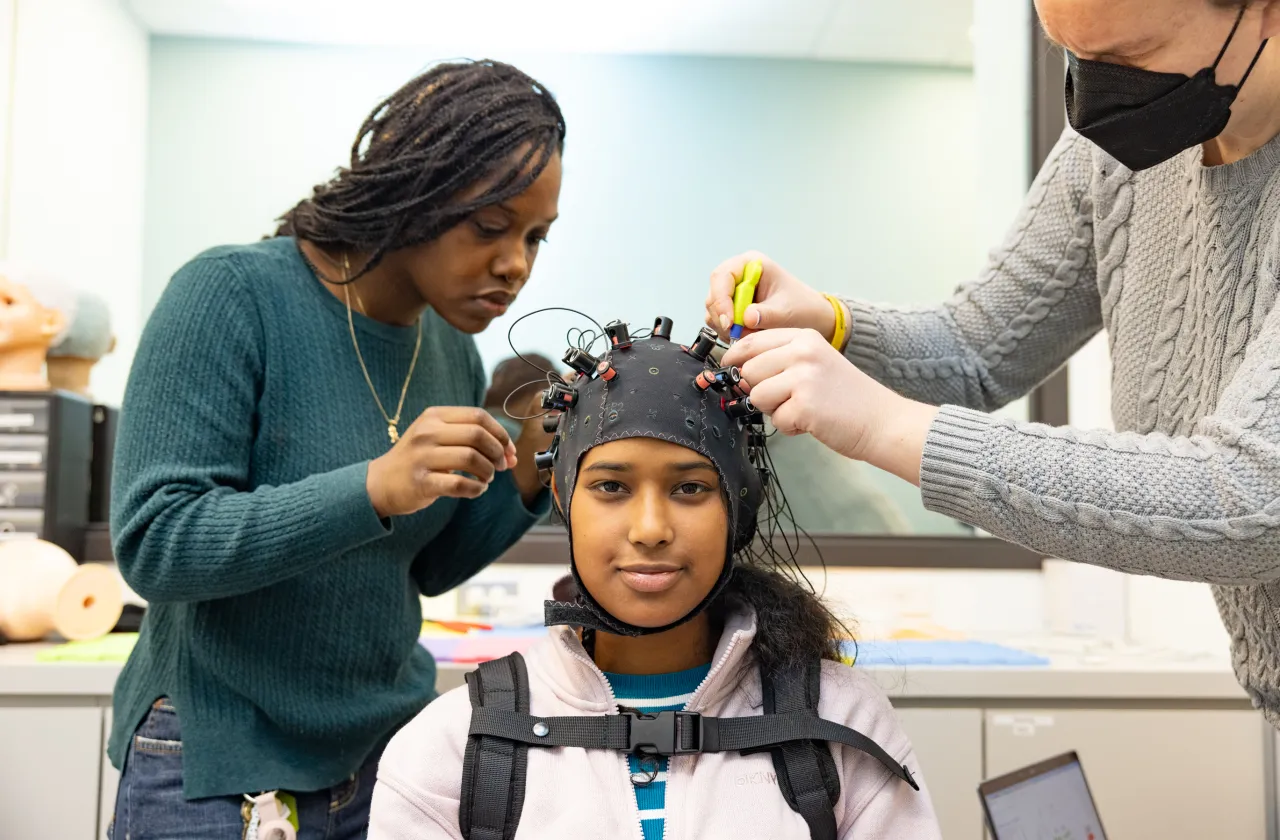 Student researcher Kris Cheaye and lab manager Maddie Scricco fit an fNIRS cap to Gloriamar Esteves’s head