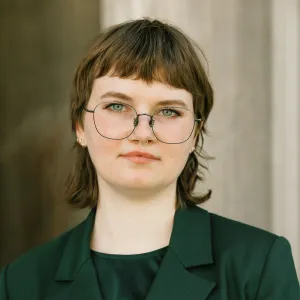 Headshot of a person with short brown hair cut into a shag style with micro-bangs, light blue eyes, and thin dark round wire-rimmed glasses. They wear a septum ring, subtle gold earrings, and a dark green blazer against a neutral, warm-toned architectural background.