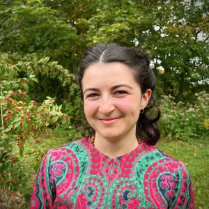 Headshot of Em Freedman smiling outdoors in a colorful pink and green paisley shirt with lush greenery in the background.