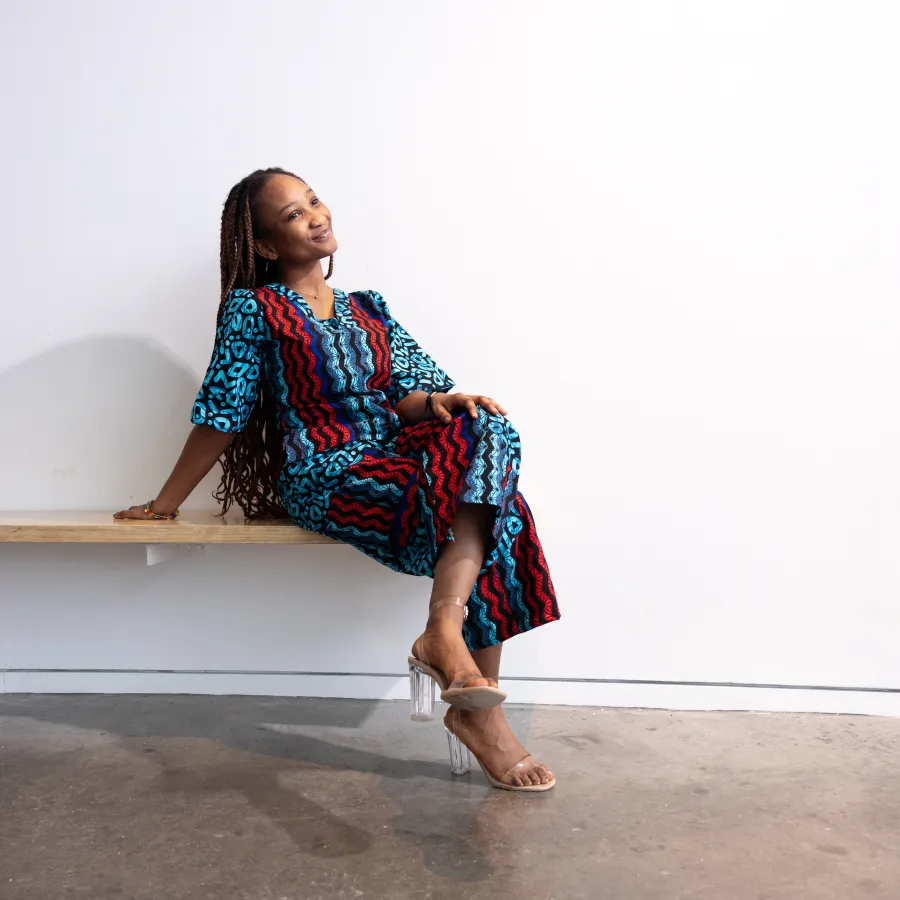 Adwoa Pokuaa Boakye sits on a bench against a white wall while wearing a red and blue printed outfit.