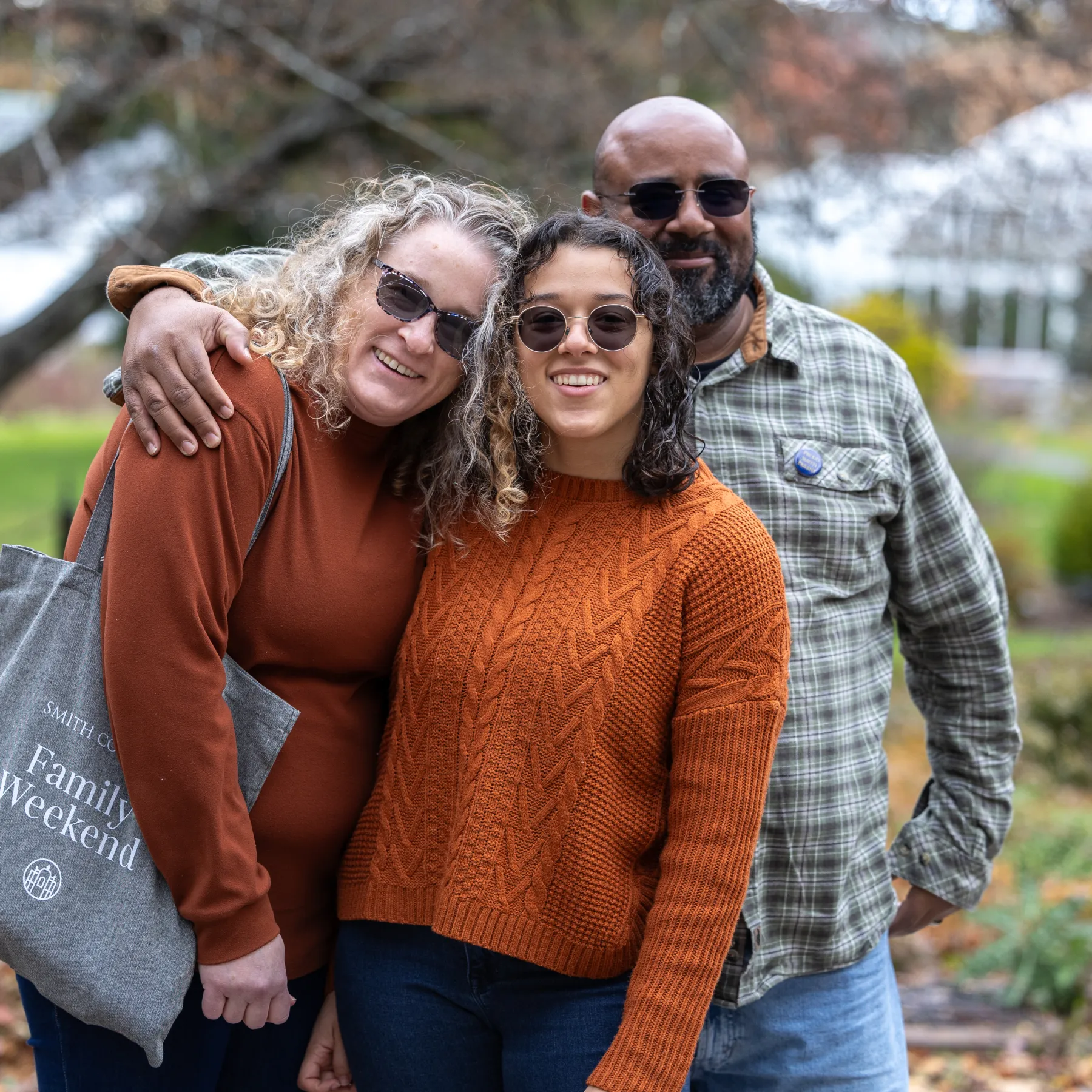 A student and parents smile at the camera. The mom is wearing a Smith College Family Weekend tote bag.