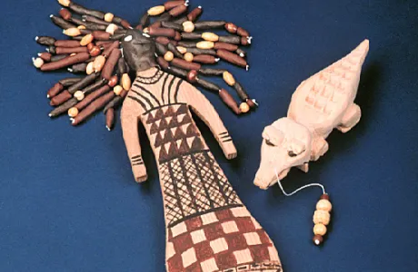 Wooden doll with bead hair and a dress with geometric patterns lies next to a carved crocodile