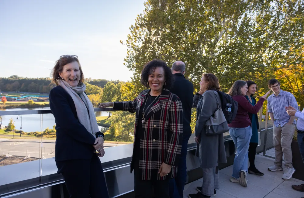 Two people smile while looking over the rooftop balcony at Kathleen McCartney Hall. In the distance are the athletic fields.