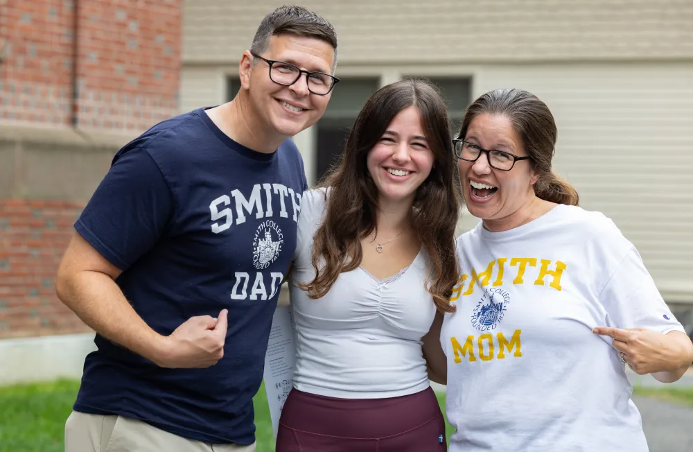 A student smiles with her parents, one is wearing a Smith Dad T-shirt and the other a Smith Mom T-shirt