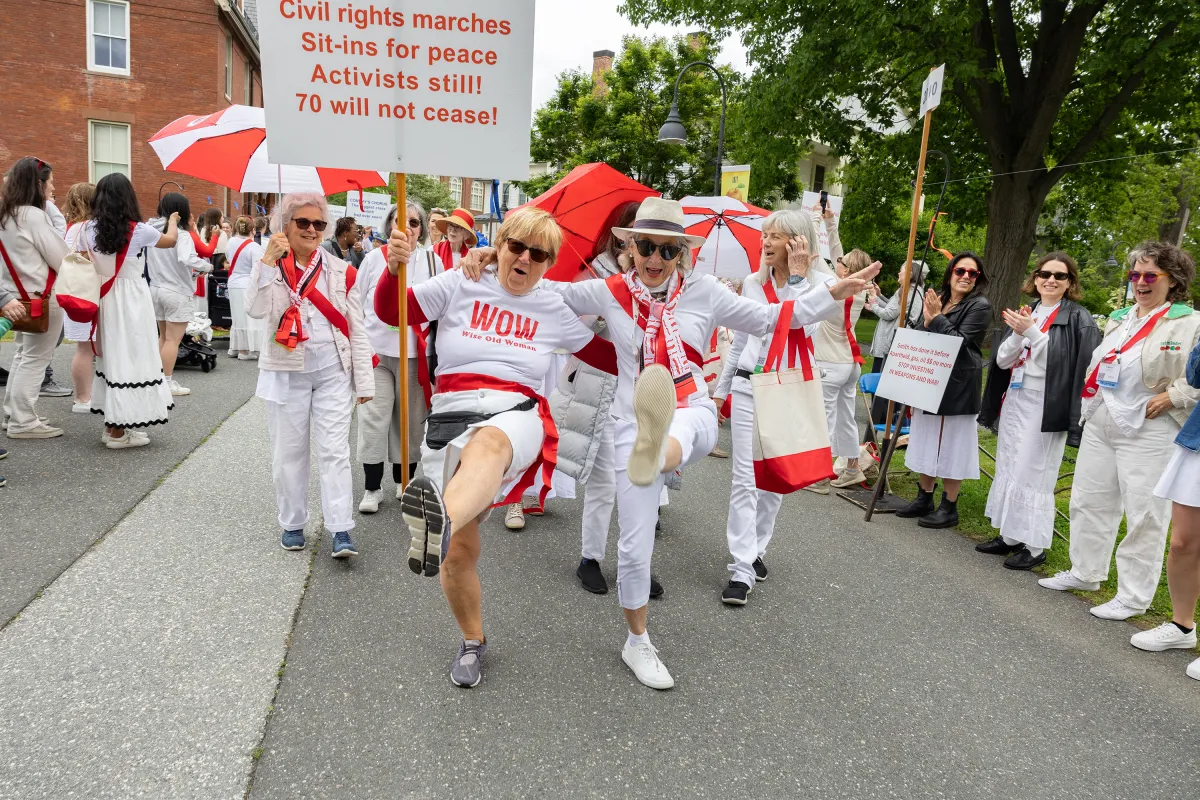 Alums from the class of 1970 wearing white and red march in the Reunion II parade. Two have their arms around one another's shoulders and they are enthusiastically kicking their legs.