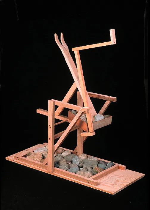A wooden abstract sculpture is set against featuring angular beams and supports. Several rocks are placed within the structure.