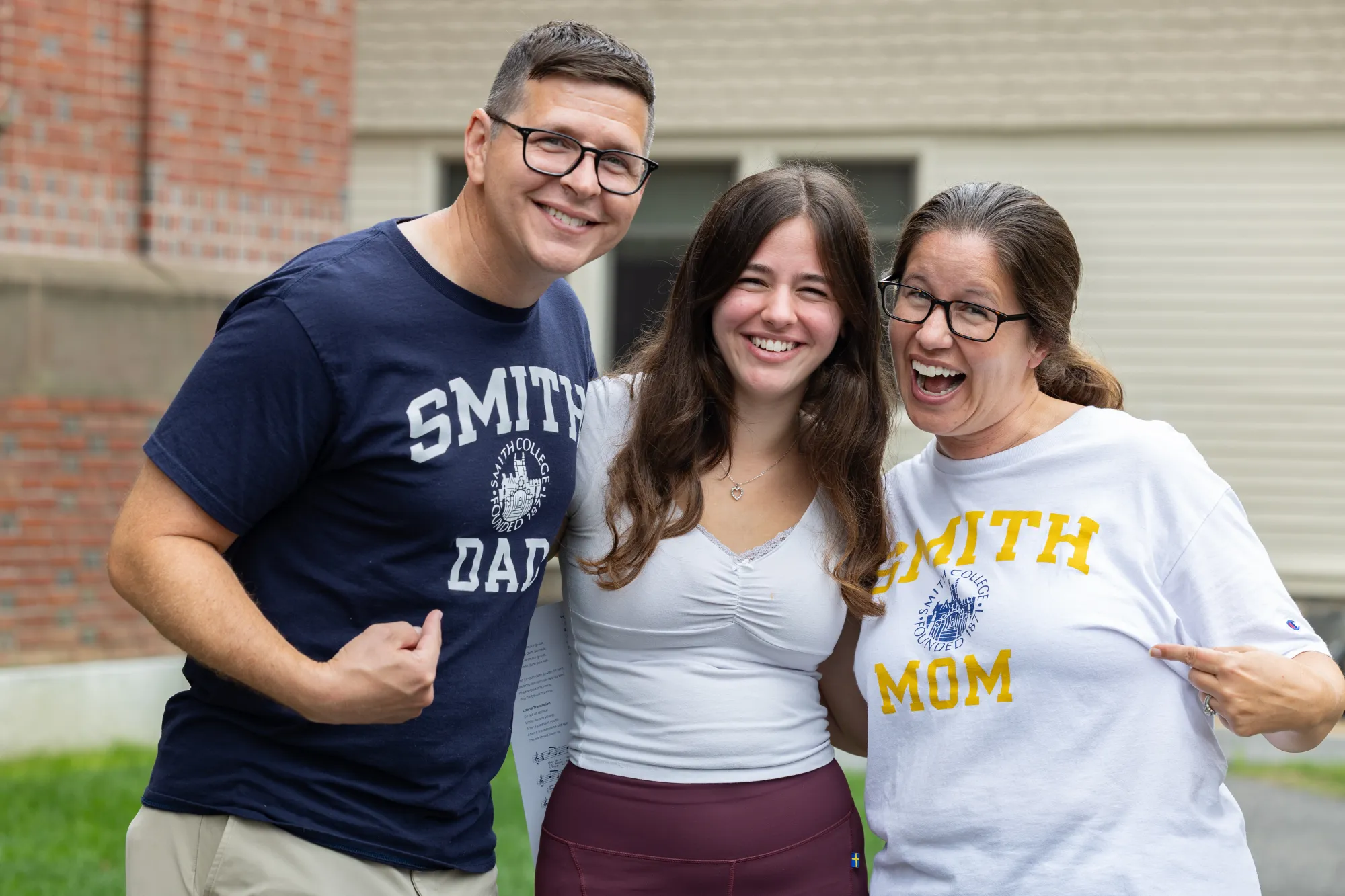 A student smiles with her parents, one is wearing a Smith Dad T-shirt and the other a Smith Mom T-shirt
