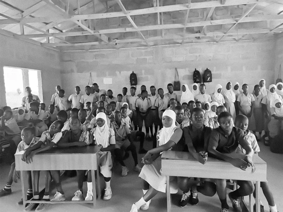 A large group of children sits, some at desks, and stands gathered inside of a building.